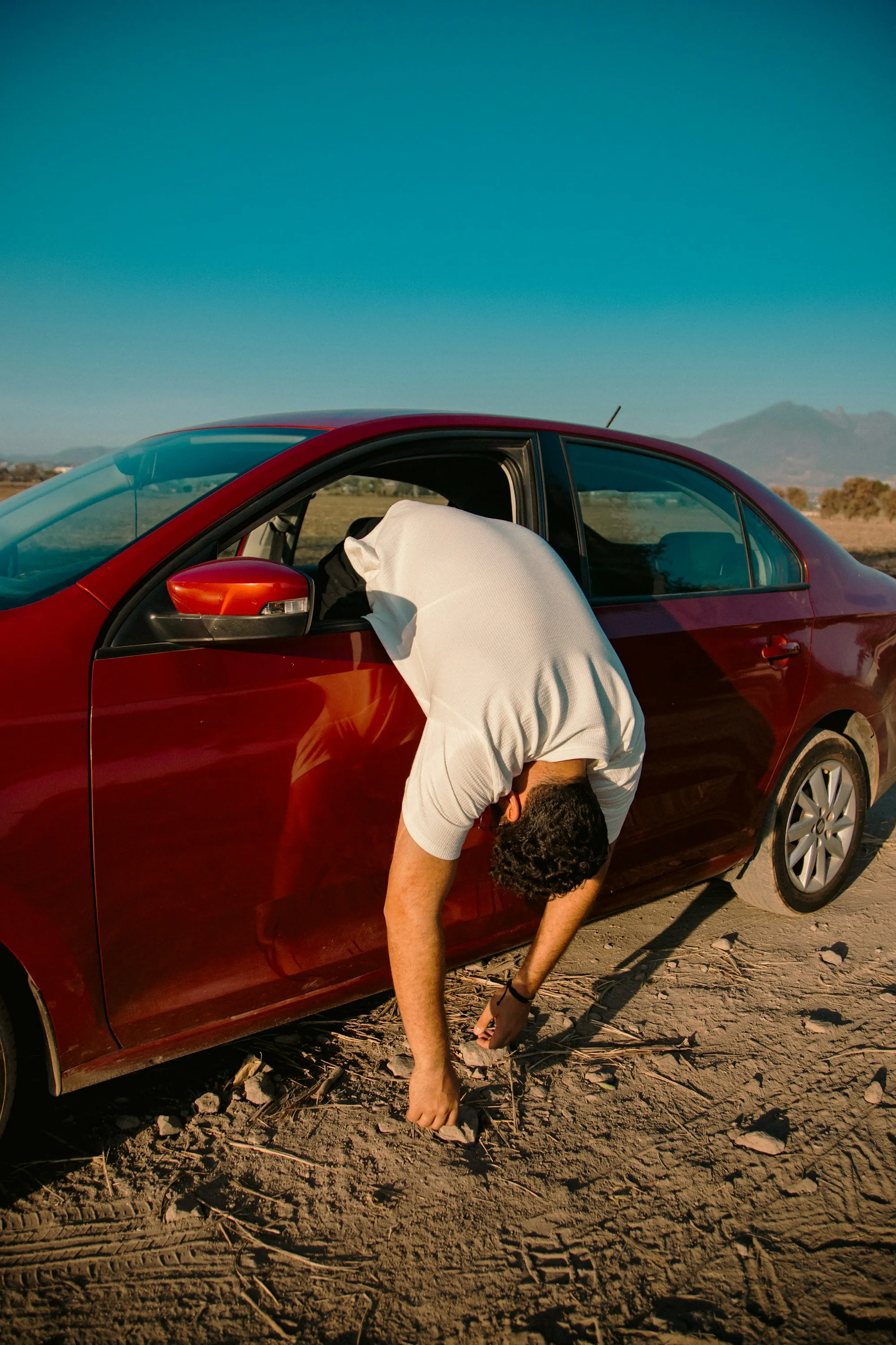 Man checking the side of a red car parked on a dirt road